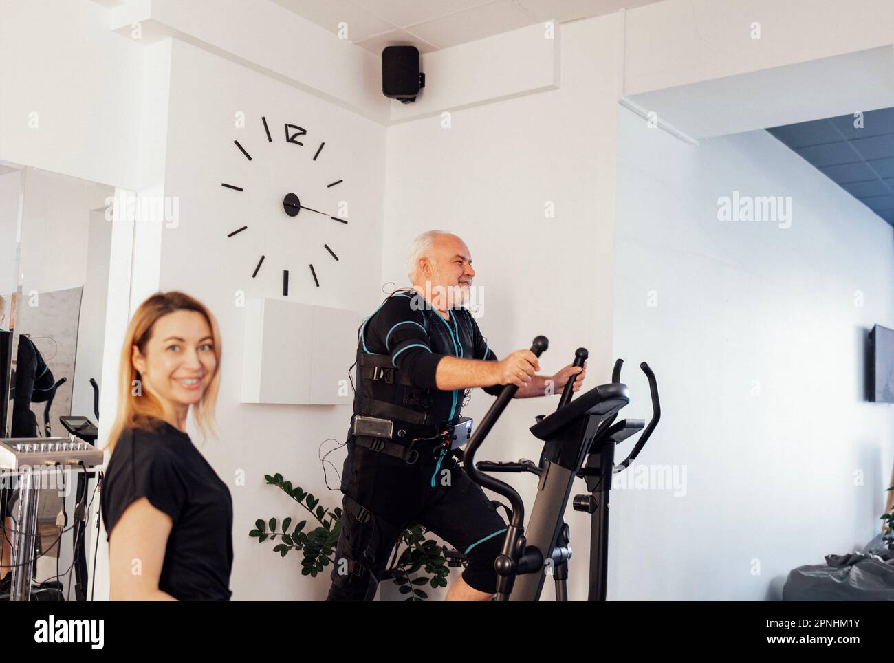 Personal trainer femminile con il suo cliente in abito ems in palestra. Sport l'uomo anziano grayhaired sta esercitandosi sull'addestratore ellittico sullo sfondo. Elettrico Foto Stock