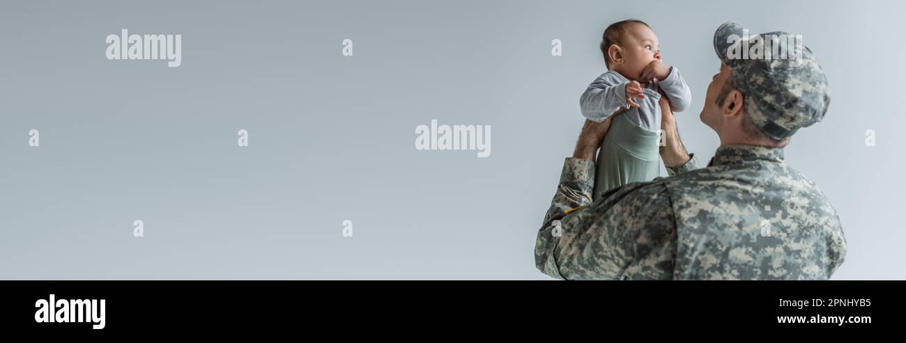 esercito soldato in uniforme e cappello tenendo bambino figlio in armi isolato su grigio, banner Foto Stock