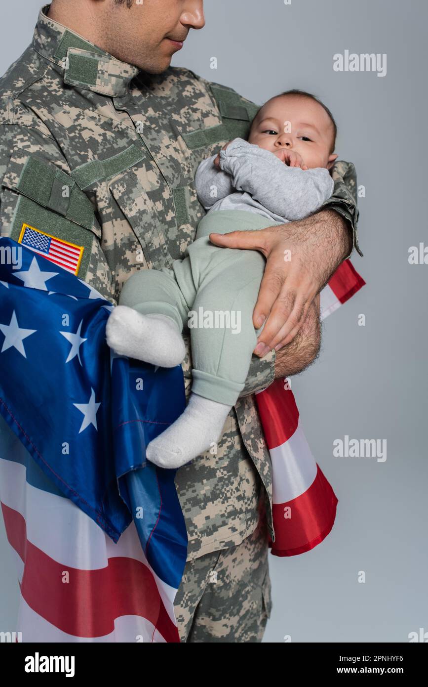 Serviceman patriottico in uniforme in piedi con bandiera d'America e tenendo il bambino in braccio sul grigio Foto Stock