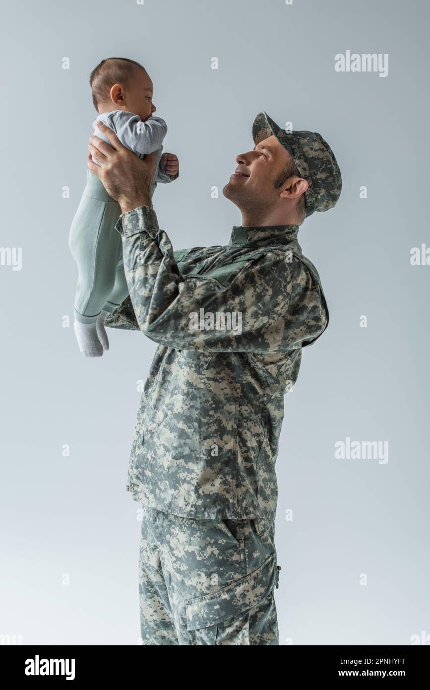 buon soldato dell'esercito in uniforme tenendo il bambino in braccio isolato sul grigio Foto Stock