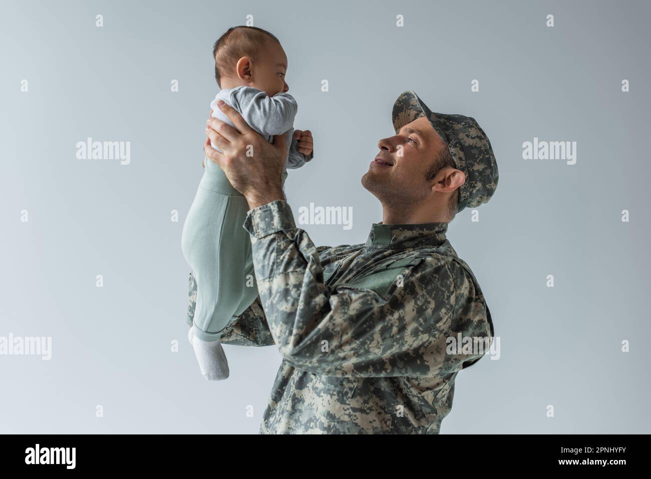buon soldato in uniforme tenendo il bambino in braccio isolato sul grigio Foto Stock