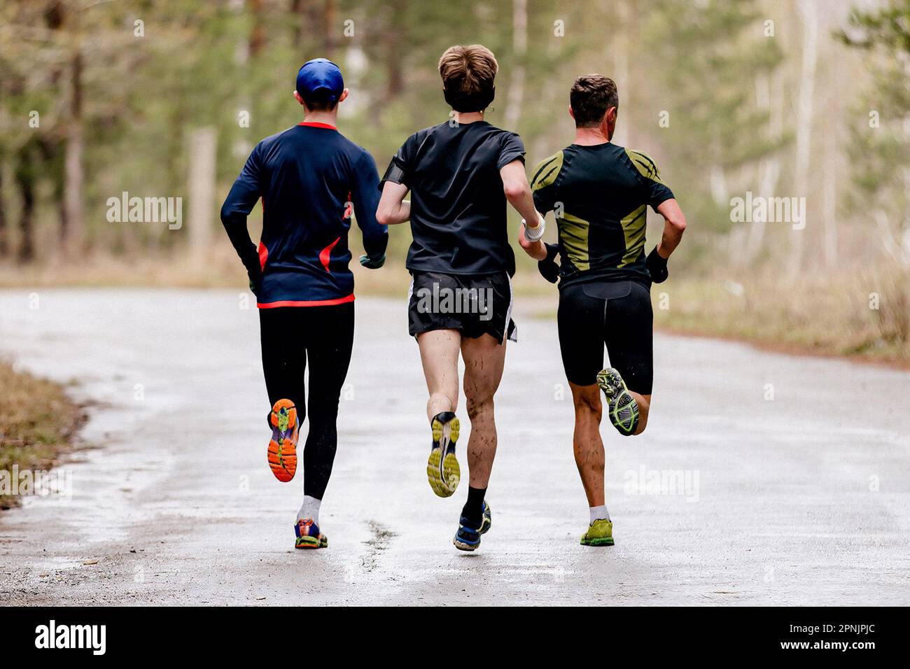 vista posteriore tre corridori maschi che corrono primavera su strada con pozzanghere, spruzzi d'acqua e sporcizia sui piedi Foto Stock