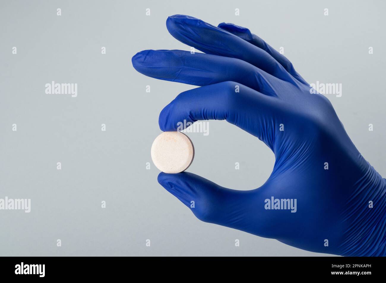 Mano maschile in un guanto medico blu che tiene una pillola bianca su uno sfondo grigio. Primo piano Foto Stock