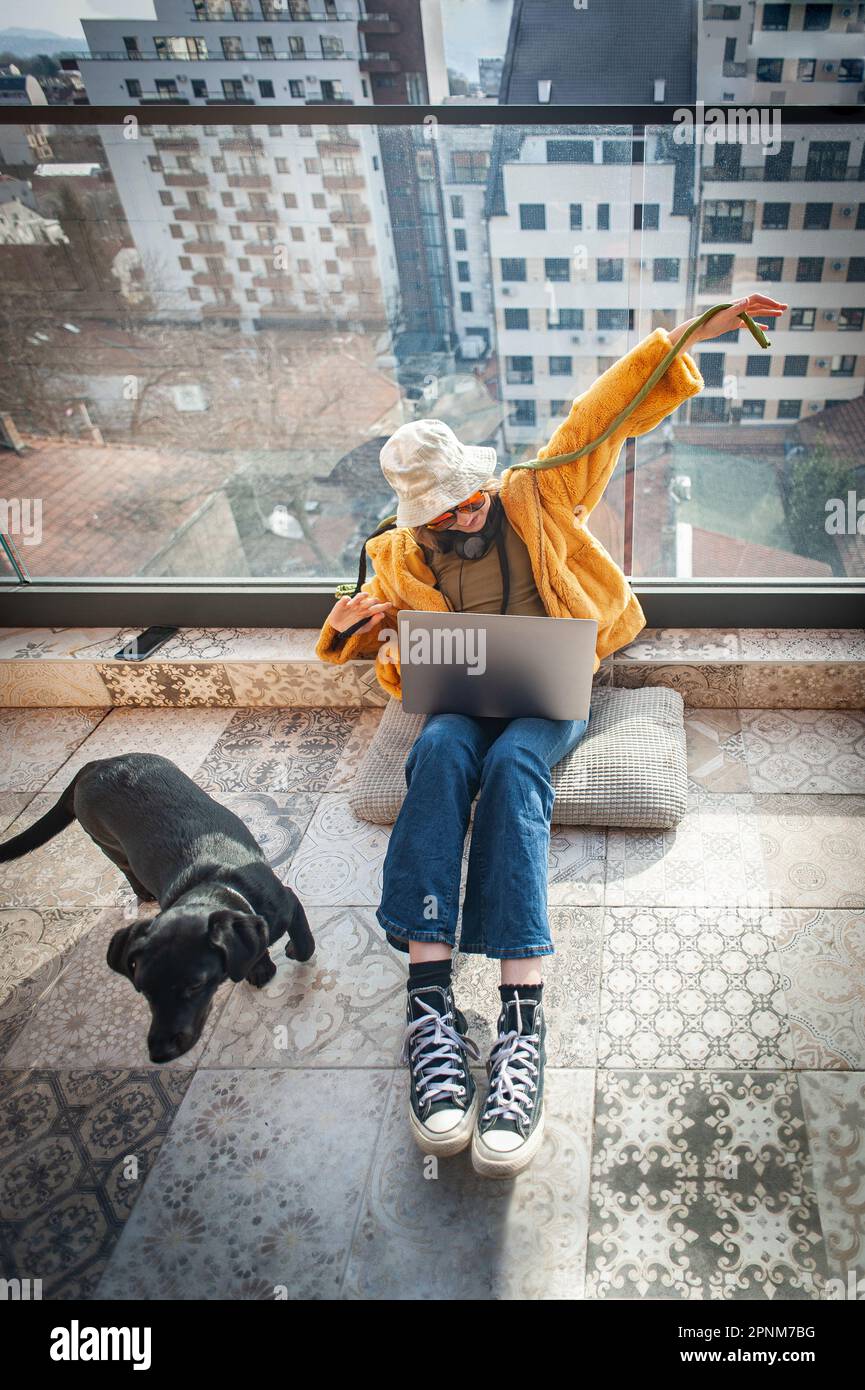 Copia spazio. Ragazza adolescente Hippster seduta sul terrazzo, allegro spalmare le braccia, utilizzando un laptop in grembo, con vista sulla città e il cielo. Giovane felice Foto Stock