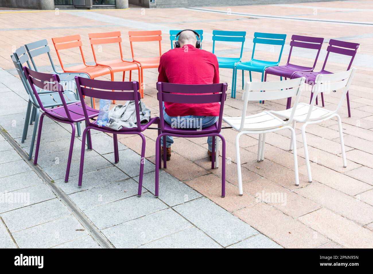 Uomo seduto in un cerchio di sedie multicolore e indossando cuffie. Foto Stock