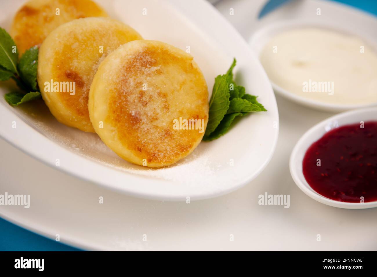 Frittelle di formaggio ricotta rotonda in un piatto bianco su sfondo blu. Cibo prima colazione. Cibo sano. Salsa di panna acida e menta. Primo piano vista dall'alto. Pasto dessert. Frittelle di ricotta al formaggio ricotta. Foto Stock
