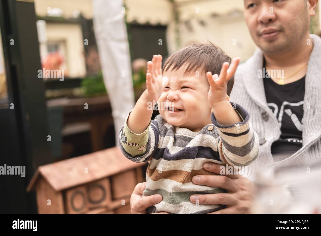 Bambino maschio sorridente alzando le mani nell'eccitamento seduto sulle gambe del suo padre, il papà sta tenendo il bambino con le sue mani. All'aperto in una birreria all'aperto in PU Foto Stock