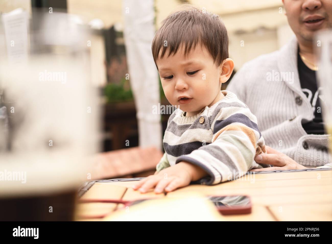Bambino maschio con sguardo serio che tocca il tavolo di legno con la mano seduta sulle gambe del padre ad un tavolo in un giardino della birra in un pub a Edimburgo, Scot Foto Stock