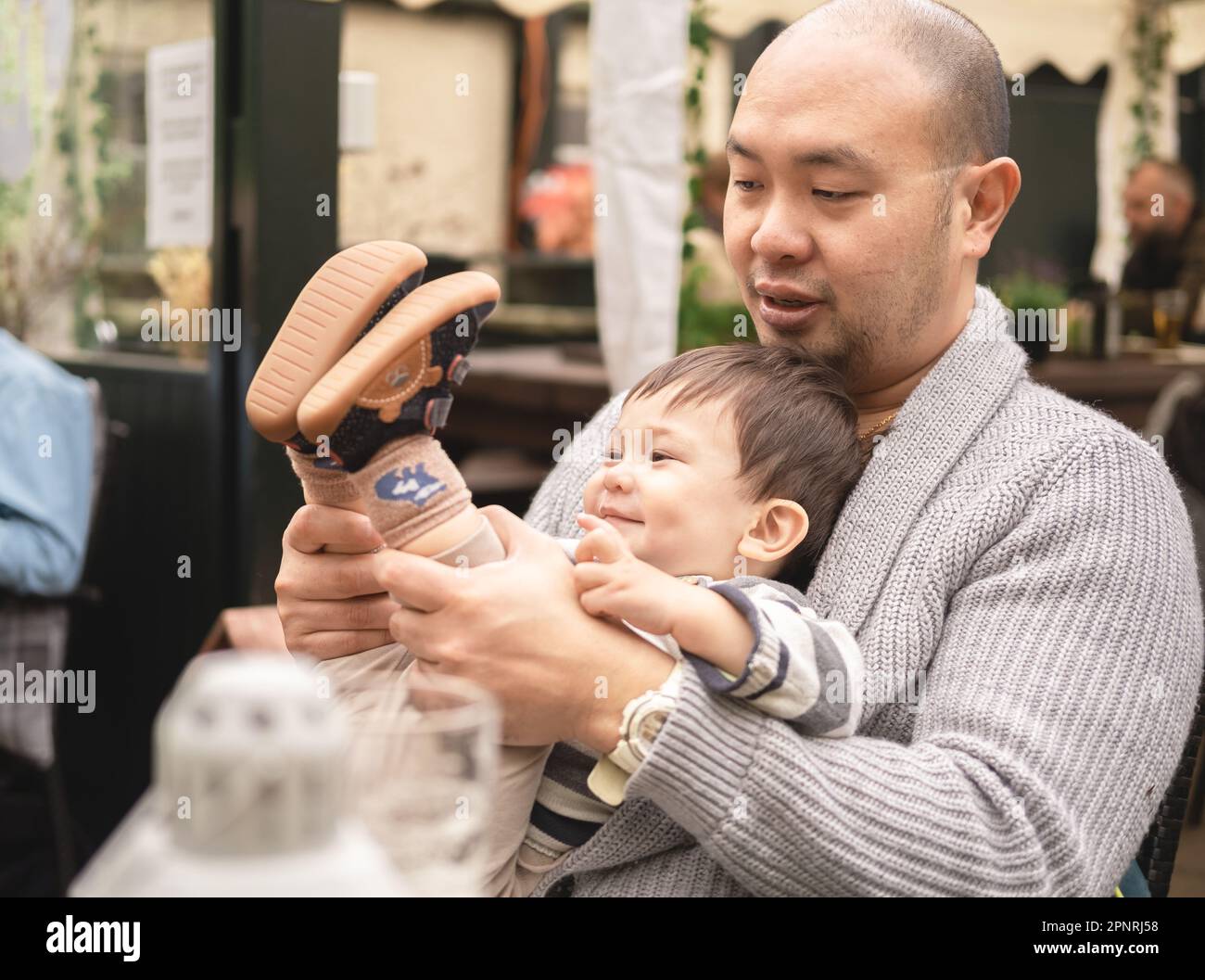 Flessibile bambino maschio sorridente seduto sulle gambe del papà tenendo le gambe del figlio, bambino che gioca con il padre in birreria all'aperto al pub. Stile di vita gioioso. H Foto Stock