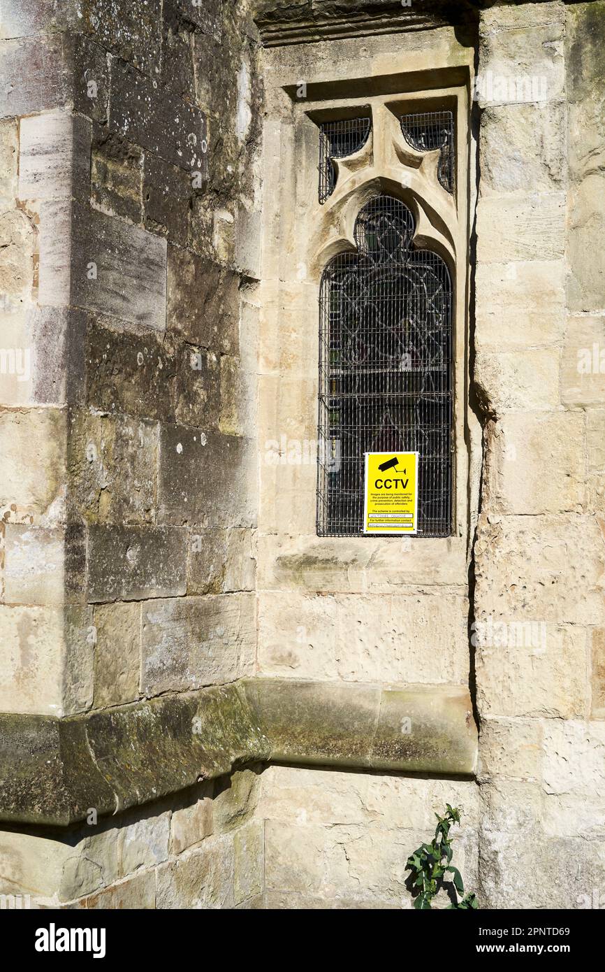 Avviso CCTV giallo visualizzato nella finestra con piombo di una vecchia chiesa in pietra protetta da una griglia metallica Foto Stock