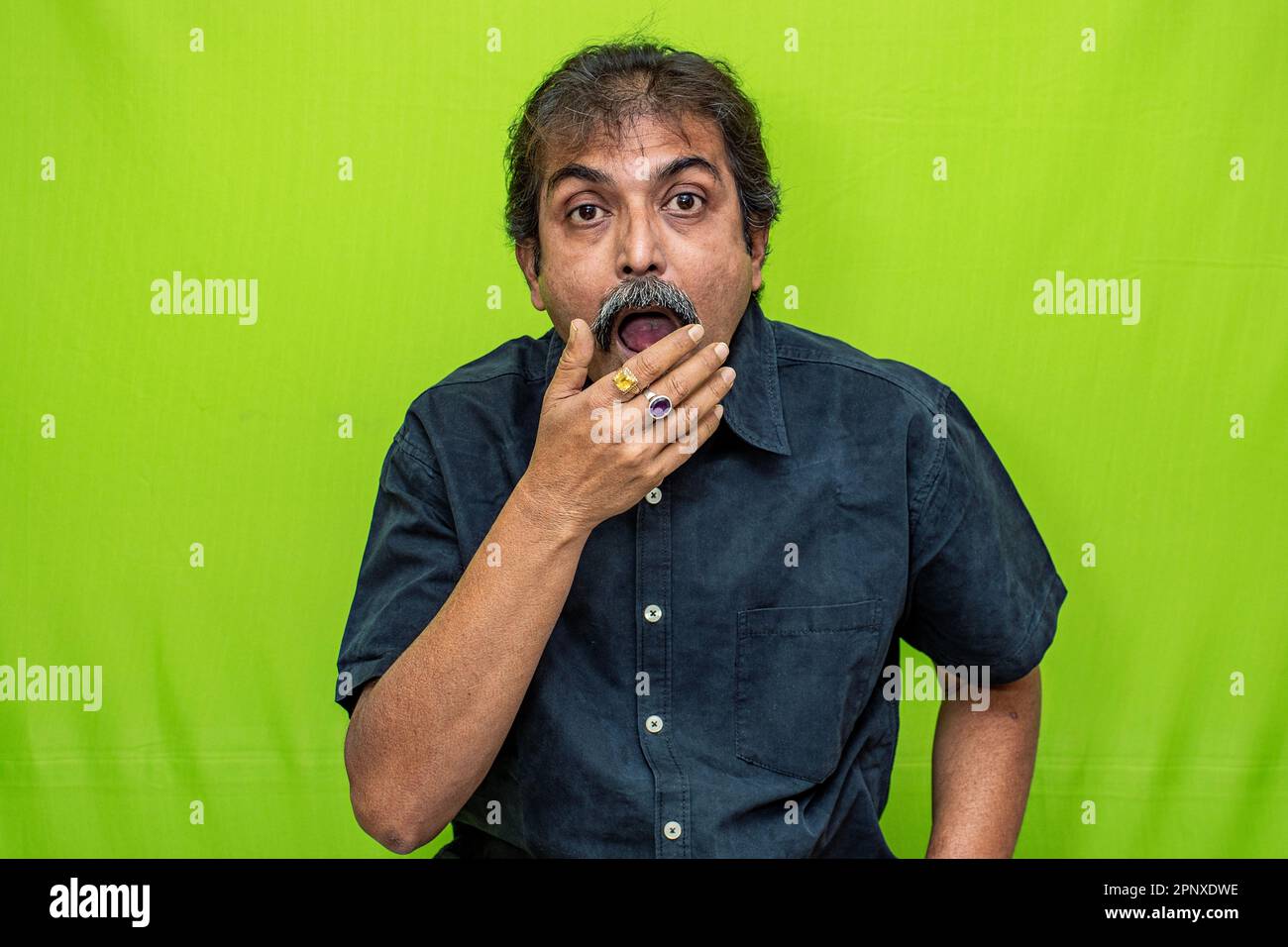 Un uomo d'affari ben vestito in una camicia nera è visto in piedi contro uno schermo verde, con la mano destra sulla sua bocca in un'espressione sorpresa Foto Stock