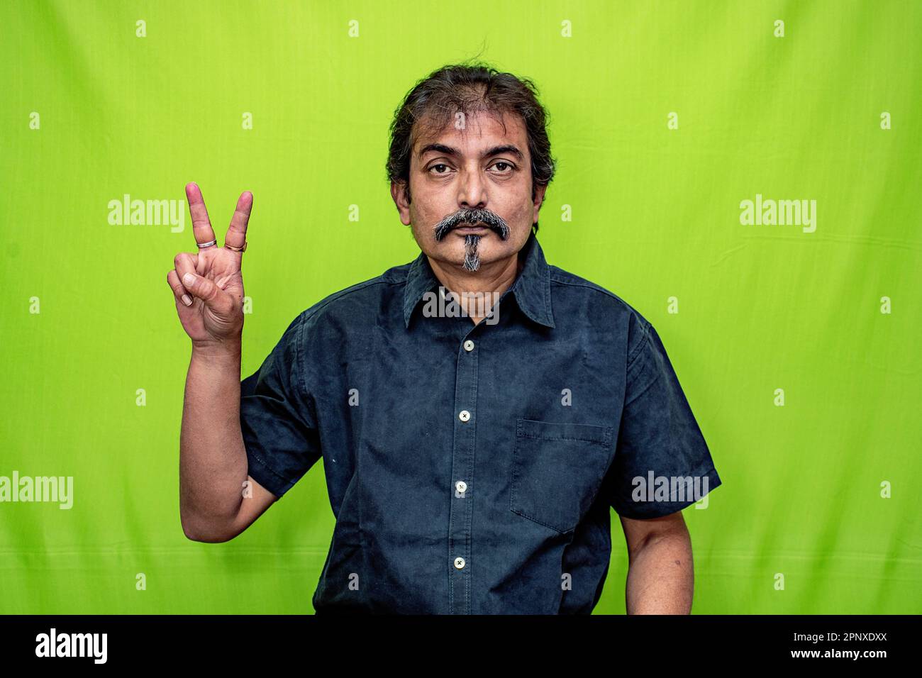 Un uomo aziendale molto intelligente in camicia nera sta mostrando 'due' con le dita della sua mano destra, in piedi contro uno sfondo verde schermo Foto Stock