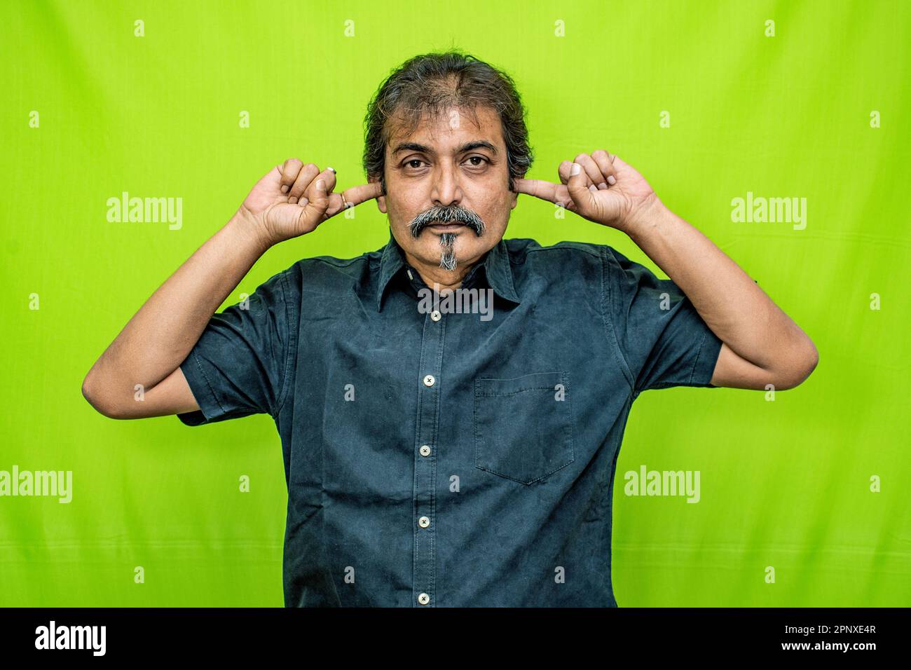 Un uomo d'affari ben vestito che indossa una camicia nera si trova contro uno schermo verde, coprendo entrambe le orecchie con i suoi forefingers, a indicare la sordità Foto Stock