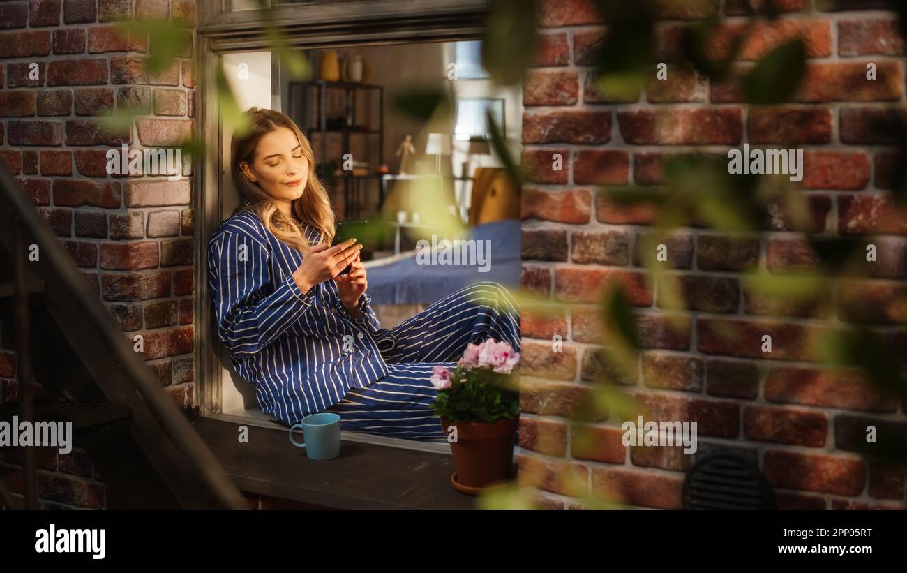 Giovane bella donna utilizzando smartphone e godendo il suo caffè mattutino mentre si siede sul suo windowsill da camera da letto. Femmina caucasica in pigiama a strisce Foto Stock