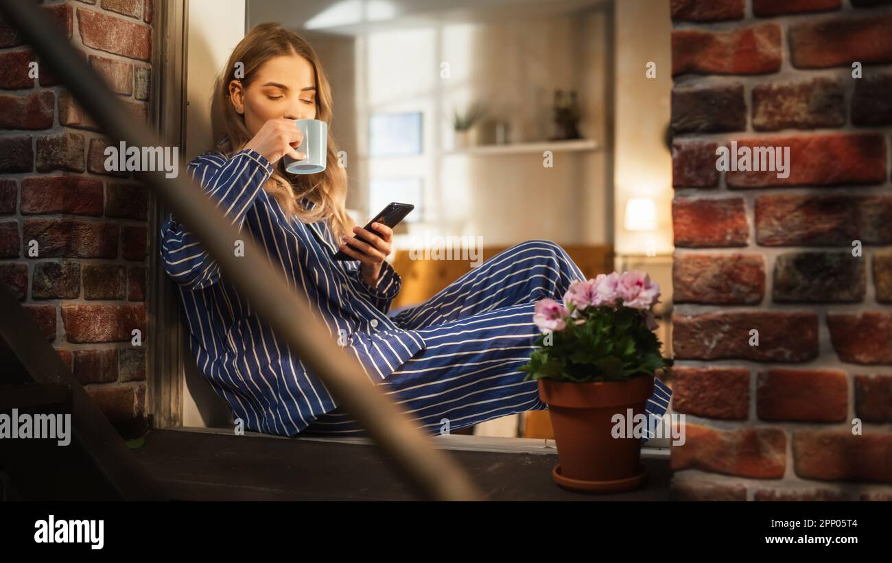 Giovane bella donna utilizzando smartphone e godendo il suo caffè mattutino mentre si siede sul suo windowsill da camera da letto. Femmina caucasica in pigiama a strisce Foto Stock