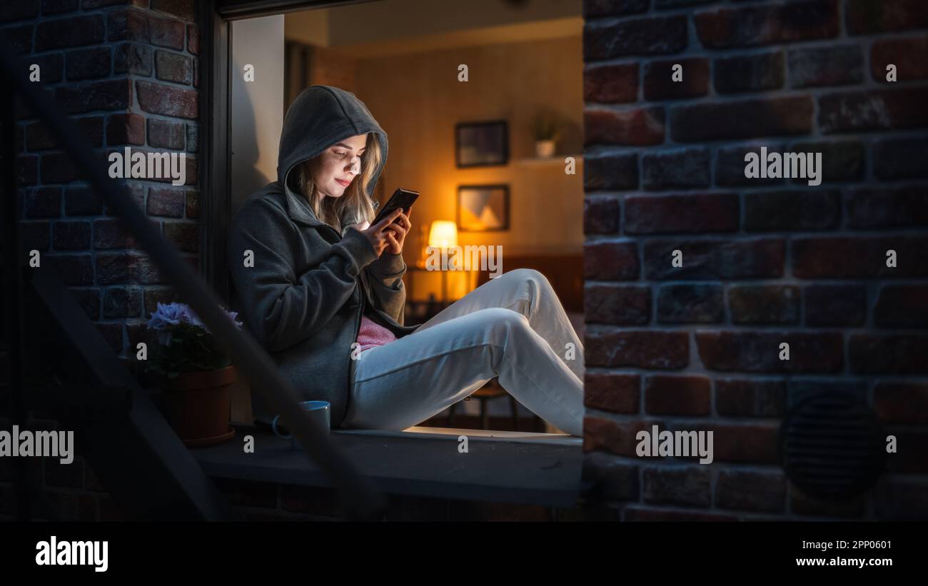 Donna bianca che indossa una felpa con cappuccio con il suo smartphone mentre si siede sul davanzale di notte. Giocatore femminile che gioca giochi addictive del telefono cellulare invece Foto Stock