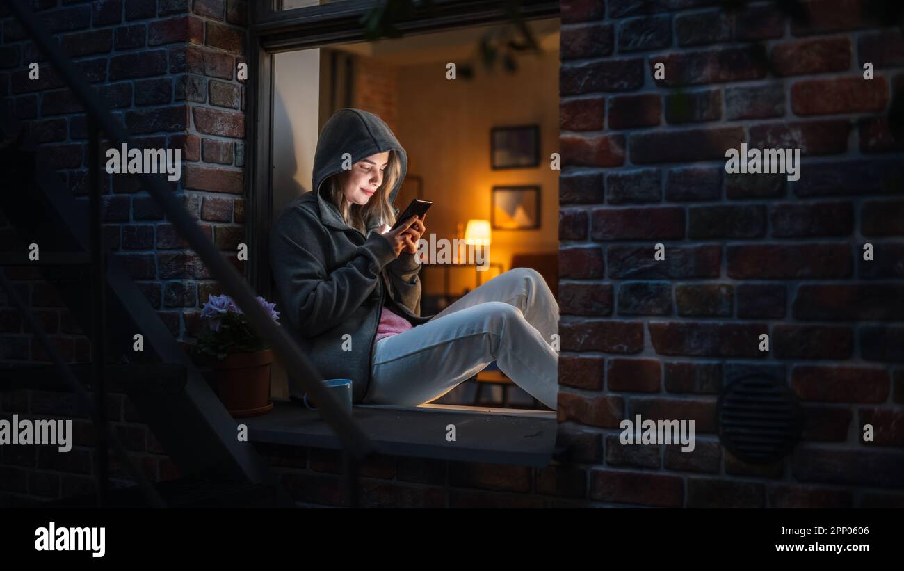 Donna bianca che indossa una felpa con cappuccio con il suo smartphone mentre si siede sul davanzale di notte. Giocatore femminile che gioca giochi addictive del telefono cellulare invece Foto Stock