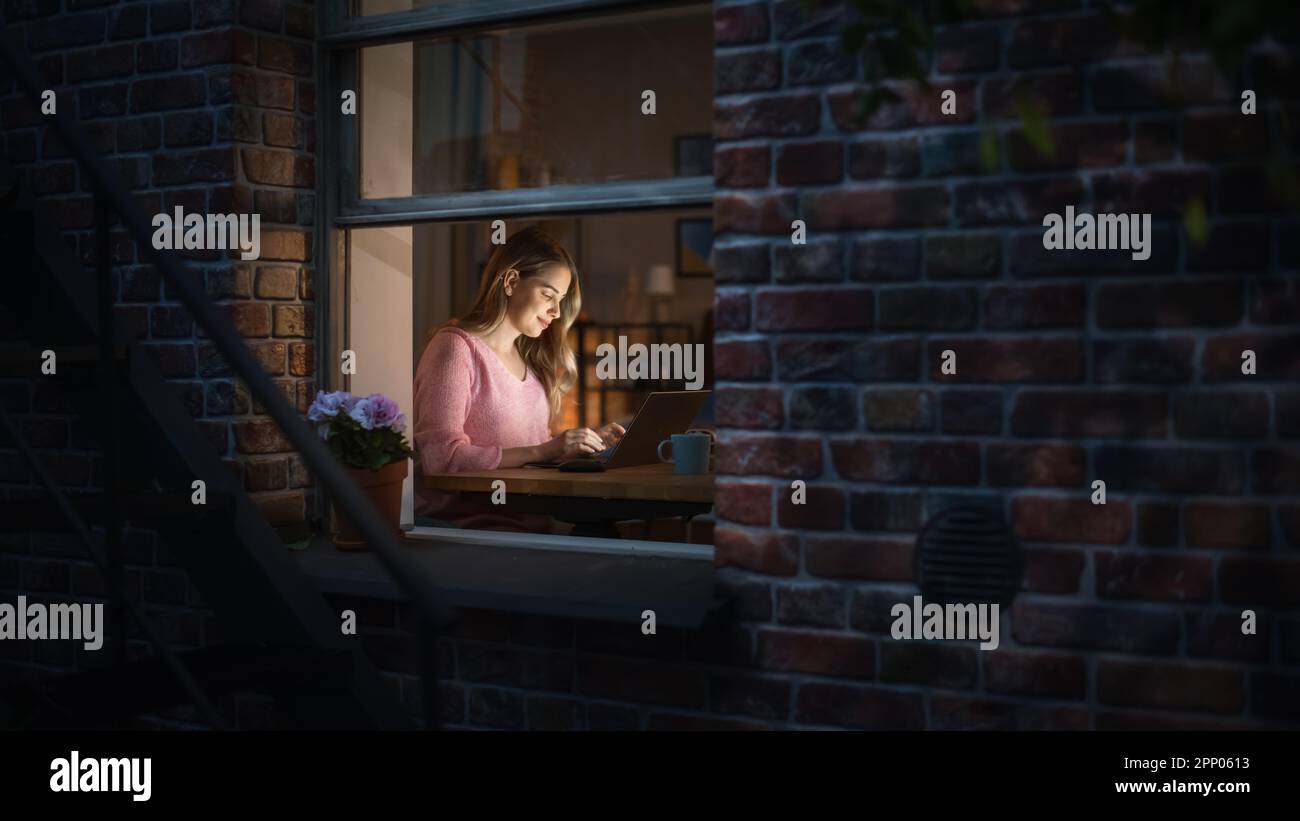 Bella donna bionda lavora su un computer portatile mentre si siede alla sua scrivania in camera da letto. Freelancer professionista femminile che lavora da casa accogliente di notte. Foto Stock