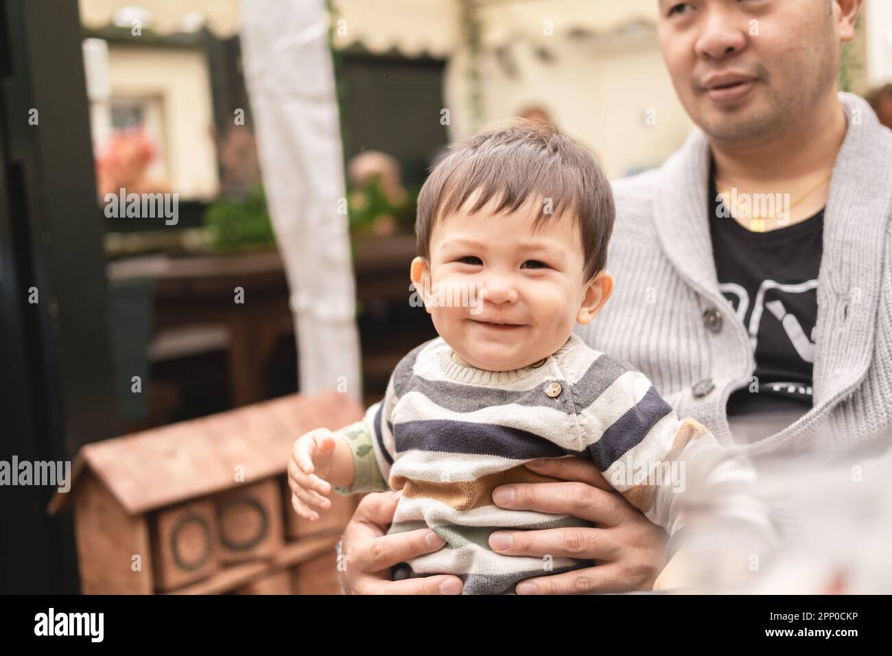 Bambino maschio sorridente seduto sulle gambe del padre guardando felicemente la macchina fotografica, papà sta tenendo il bambino con le mani. Seduti all'aperto in una birreria all'aperto in un bar Foto Stock