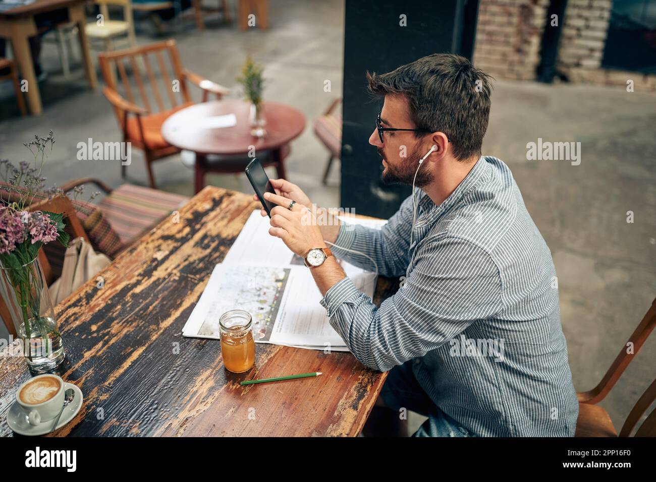 Giovane uomo seduto da solo in un bar, utilizzando lo smartphone e ascoltando le cuffie. Bell'interno di una caffetteria moderna. Foto Stock