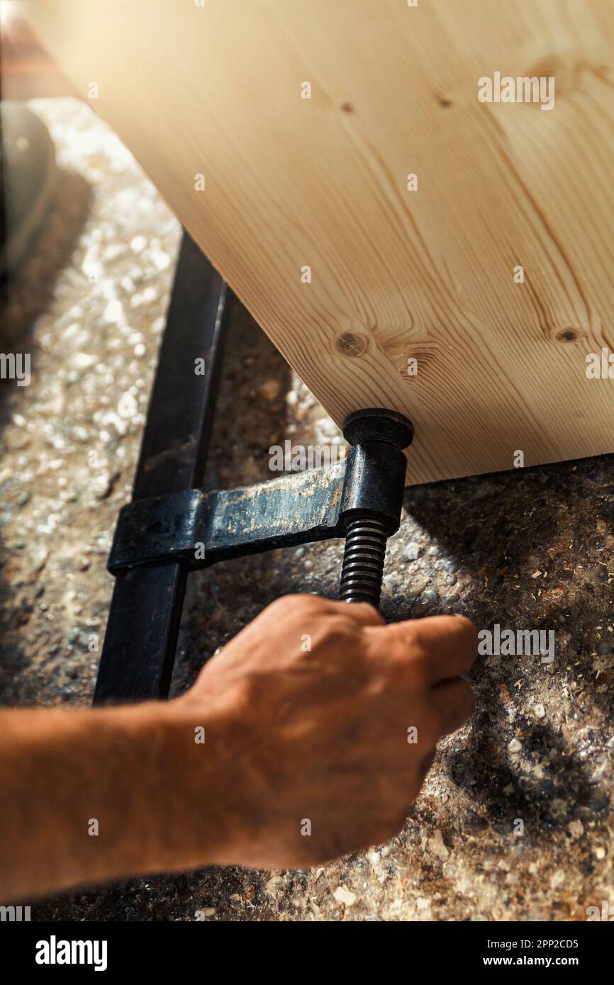 La mano del falegname fissa due parti di un mobile con un morsetto a vite in legno nel suo laboratorio artigianale, illuminato da luce solare calda che cola Foto Stock