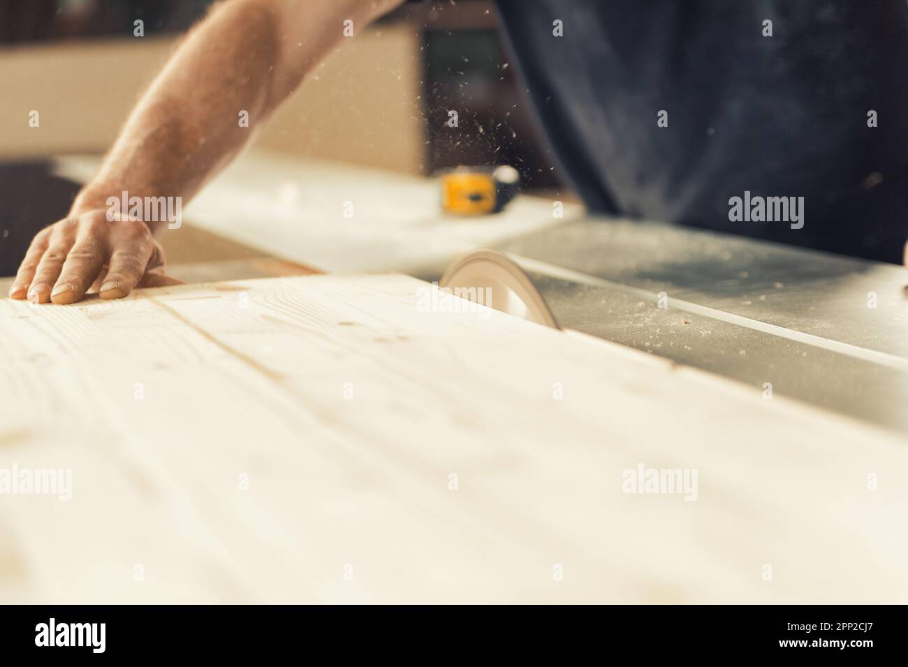Il falegname guida un asse di legno con una mano forte e sicura mentre viene tagliato da una sega circolare. Trucioli di legno e segatura volano attraverso l'aria come il Foto Stock