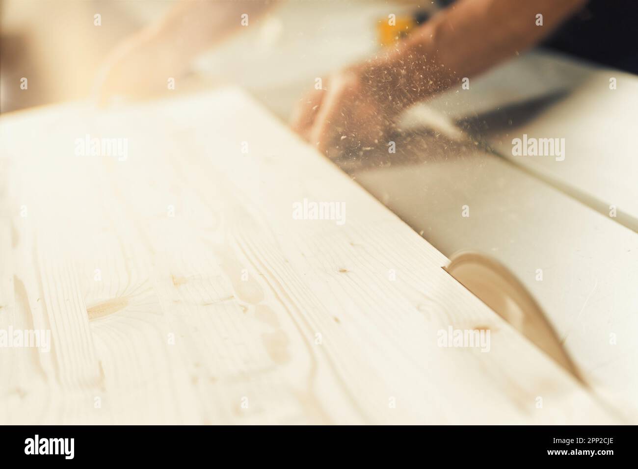 Il falegname guida un asse di legno con una mano forte e sicura mentre viene tagliato da una sega circolare. Trucioli di legno e segatura volano attraverso l'aria come il Foto Stock