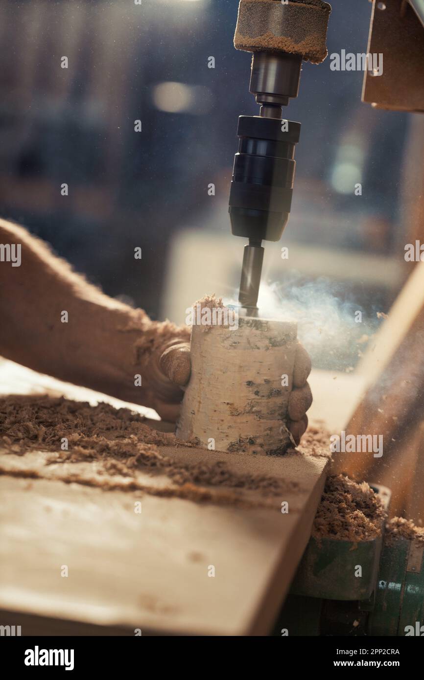 La mano forte del falegname tiene saldamente un tronco di betulla mentre si usa una pressa per trapano per intagliarlo, abbassando la leva con l'altro braccio. Le opere artigianali Foto Stock