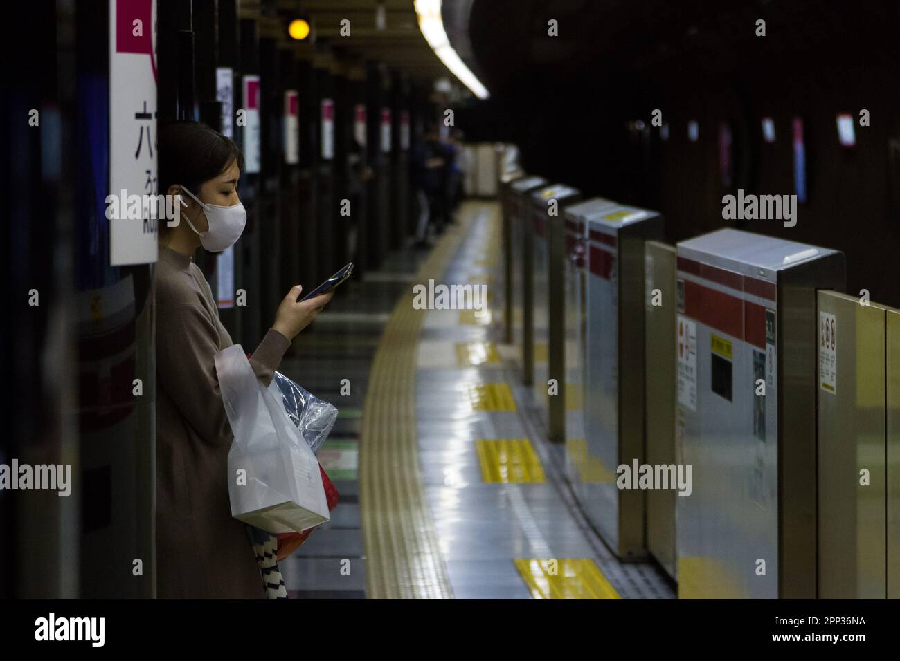 Una giovane donna giapponese, che indossa una maschera chirurgica, usa il suo smartphone mentre attende un treno alla stazione di Roppongi con barriere di sicurezza per la piattaforma Foto Stock