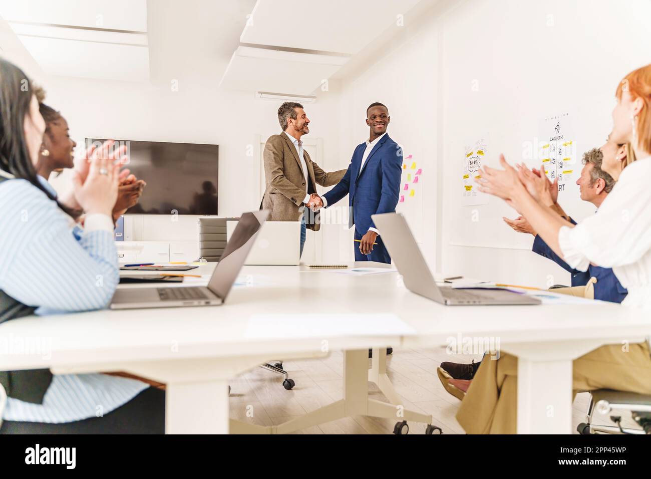 Ampio scatto di un ufficio diversificato con due persone che scuotono le mani al centro, mentre i colleghi seduti alla loro scrivania applaudono. Potrebbe significare un nuovo collab Foto Stock