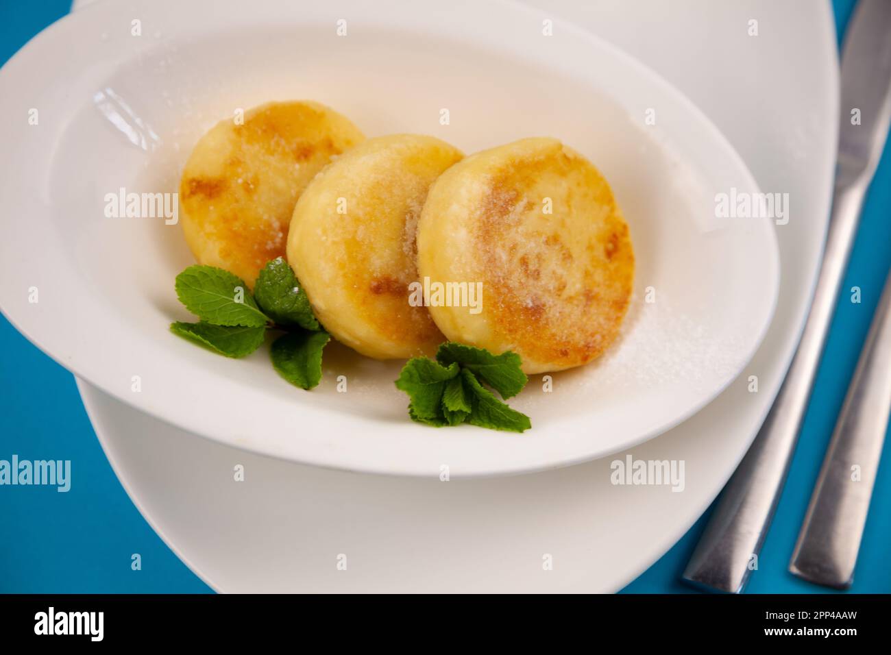 Frittelle di formaggio ricotta rotonda in un piatto bianco su sfondo blu. Cibo prima colazione. Cibo sano. Salsa di panna acida e menta. Primo piano vista dall'alto. Pasto dessert. Frittelle di ricotta al formaggio ricotta. Foto Stock