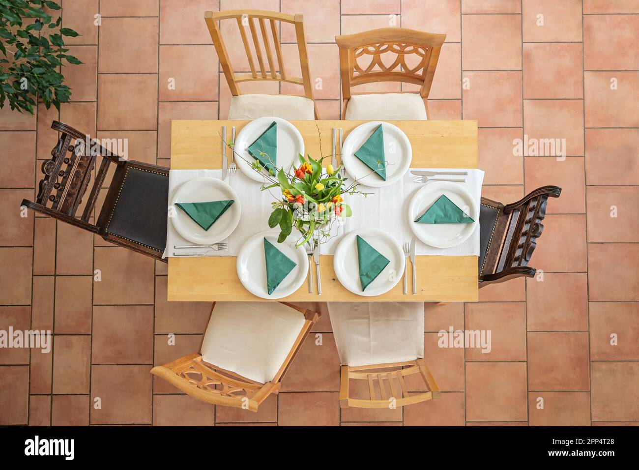 Tavolo in legno con disposizione per sei con piatti bianchi, posate, tovaglioli verdi e bouquet di fiori, diverse sedie d'epoca su terracotta piastrellata Foto Stock
