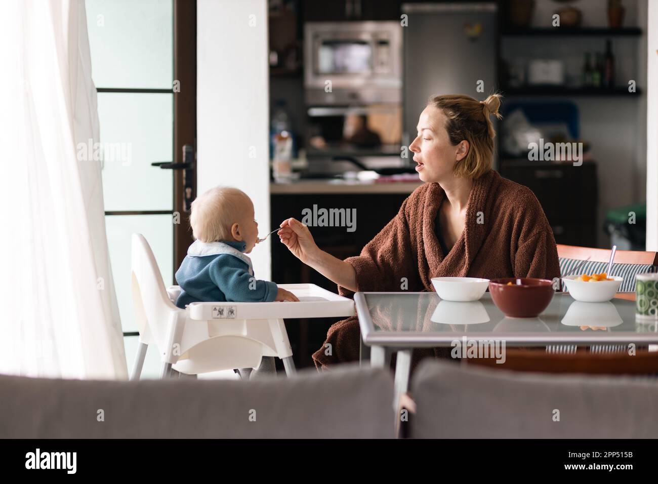 La mamma indossa il cucchiaio da bagno che allatta il bambino seduto in poltrona al tavolo da pranzo in cucina a casa la mattina. Foto Stock