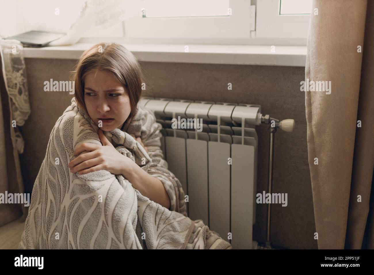 Una donna sofferente in un plaid si siede alla batteria di riscaldamento e si riscalda in casa e si congela. Foto Stock
