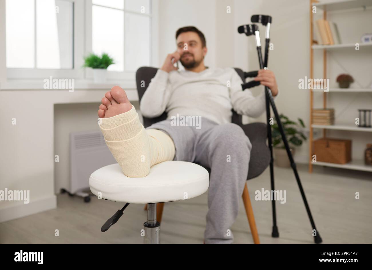 Un giovane uomo malsano è seduto a casa in sedia con gamba rotta in cast e parlando al telefono. Foto Stock