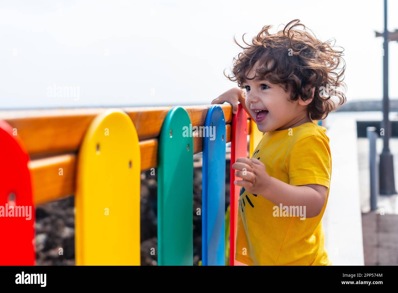 Carino ragazzino con grandi occhi e lunghi capelli sorridenti in un gioco insieme alle barriere colorate del parco Foto Stock