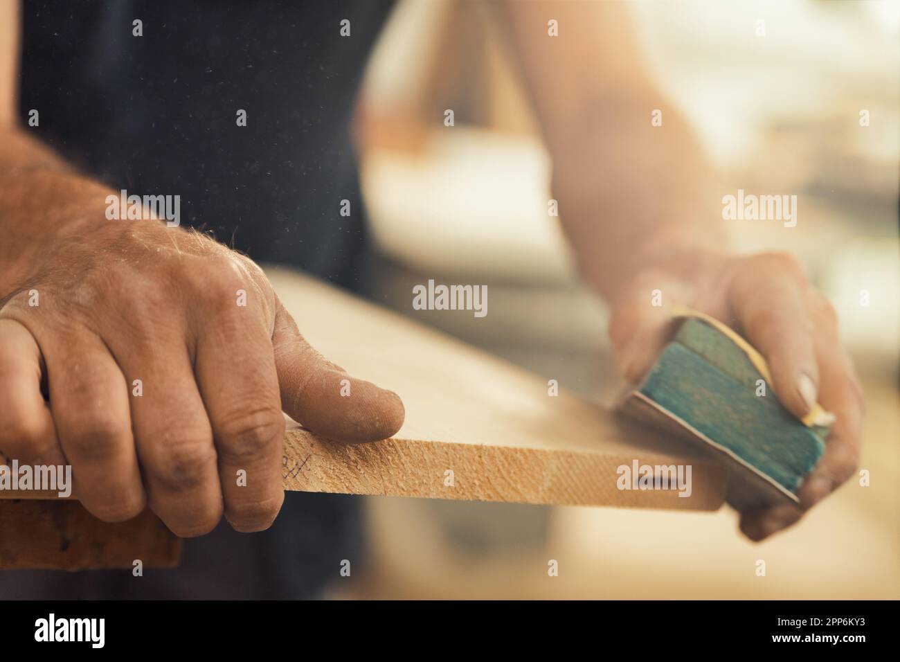 Un primo piano delle mani del falegname utilizzando carta vetrata per carteggiare manualmente un asse, appoggiandolo sulla sua superficie di lavoro. Il resto dello scatto è sfocato Foto Stock
