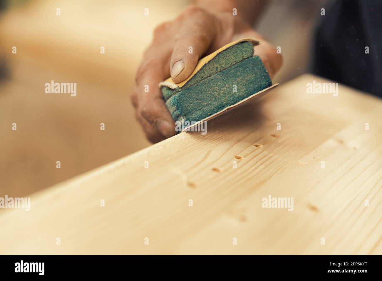 Un primo piano delle mani del falegname utilizzando carta vetrata per carteggiare manualmente un asse, appoggiandolo sulla sua superficie di lavoro. Il resto dello scatto è sfocato Foto Stock