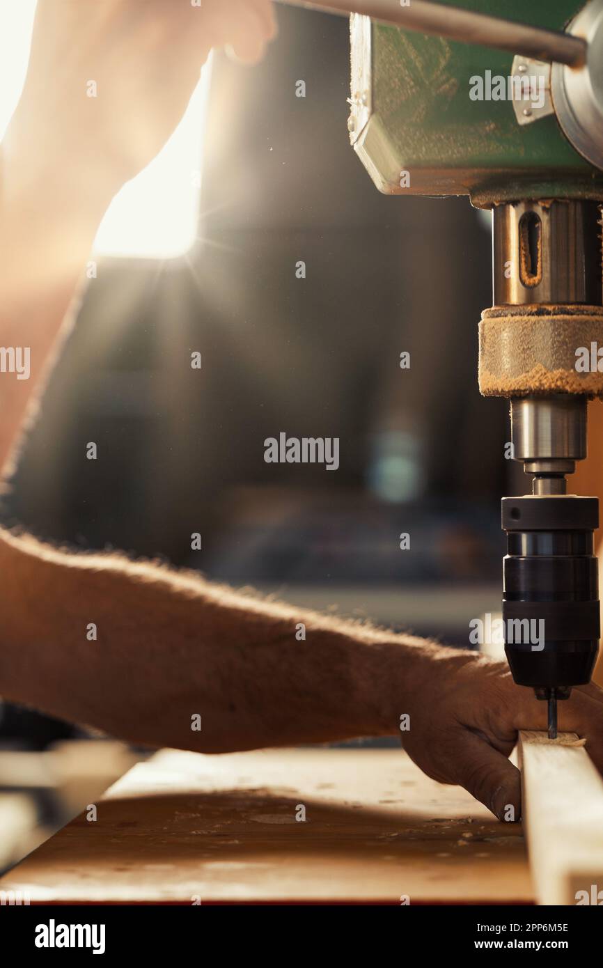 Il falegname utilizza la pressa per trapano nella sua officina. Con la mano sinistra, assumendo rischi calcolati, tiene e guida il legno, abbassando la leva con h Foto Stock