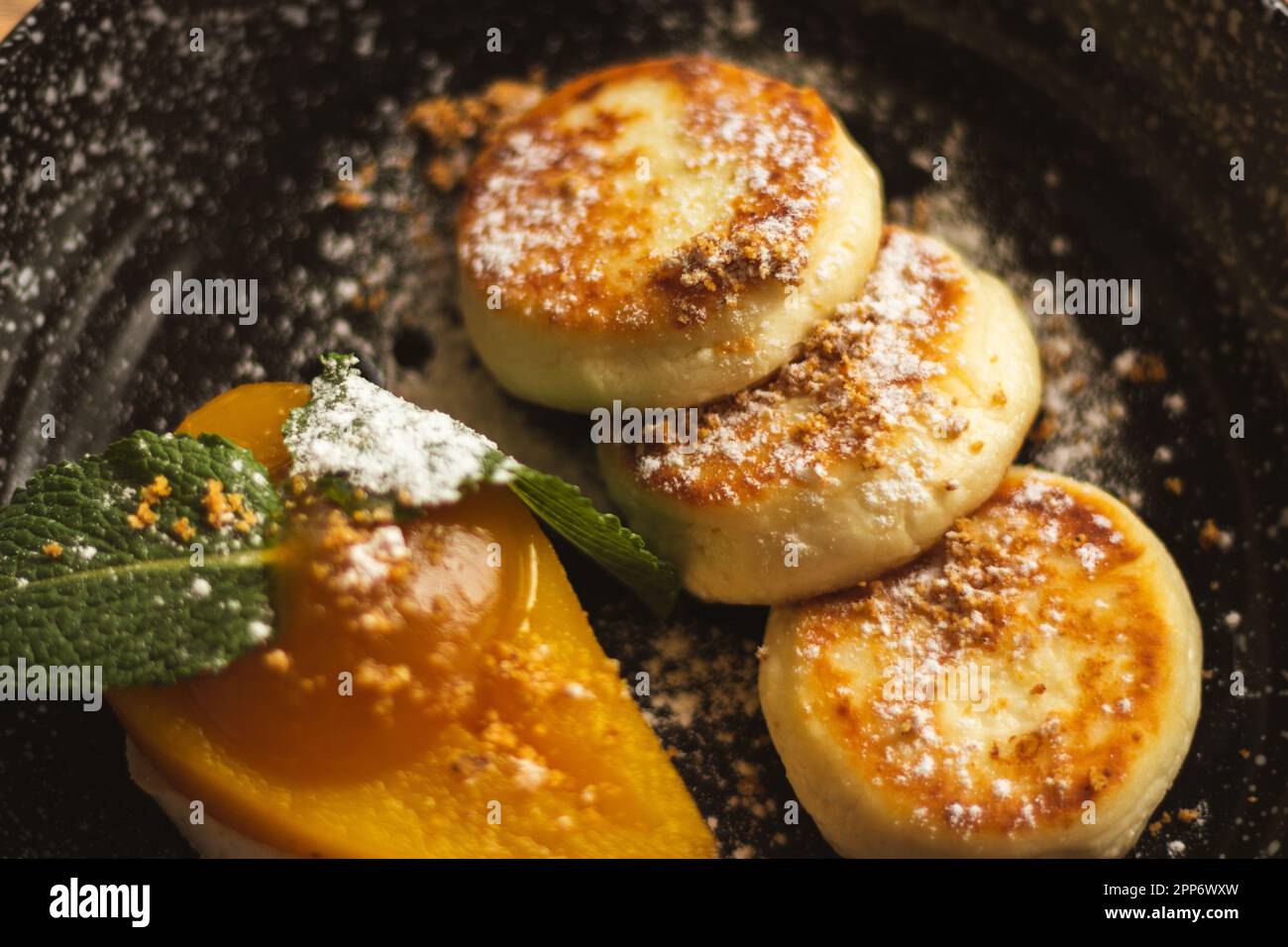 Syrniki con pera al caramello e ricotta. Frittelle cagliate su piatto nero. Colazione deliziosa. Cibo ucraino. Pera al forno con foglia di menta e frittella Foto Stock