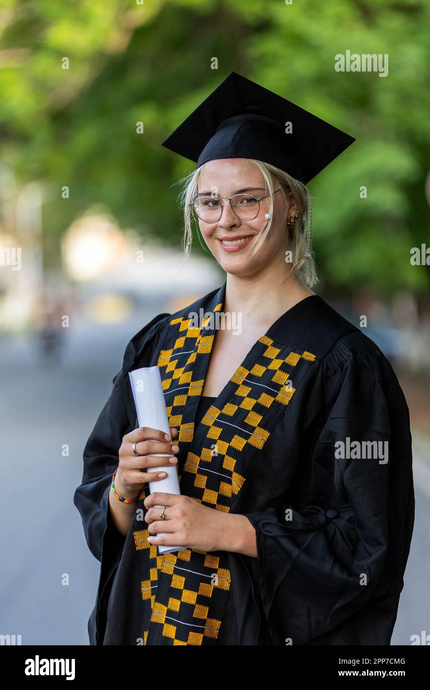Caucasian Female University Graduate indossando abito nero e berretto, tenendo il suo certificato di laurea, orgoglioso del successo accademico, e guadagnando kn Foto Stock