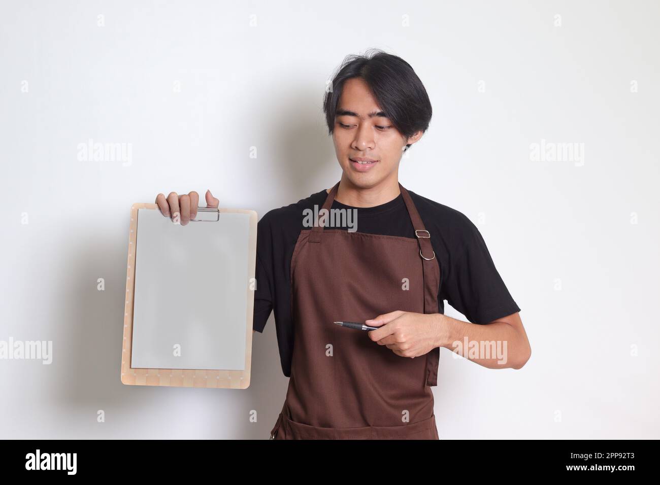 Ritratto dell'attraente barista asiatico in grembiule marrone con carta bianca del menu sulla clipboard. Immagine isolata su sfondo bianco Foto Stock