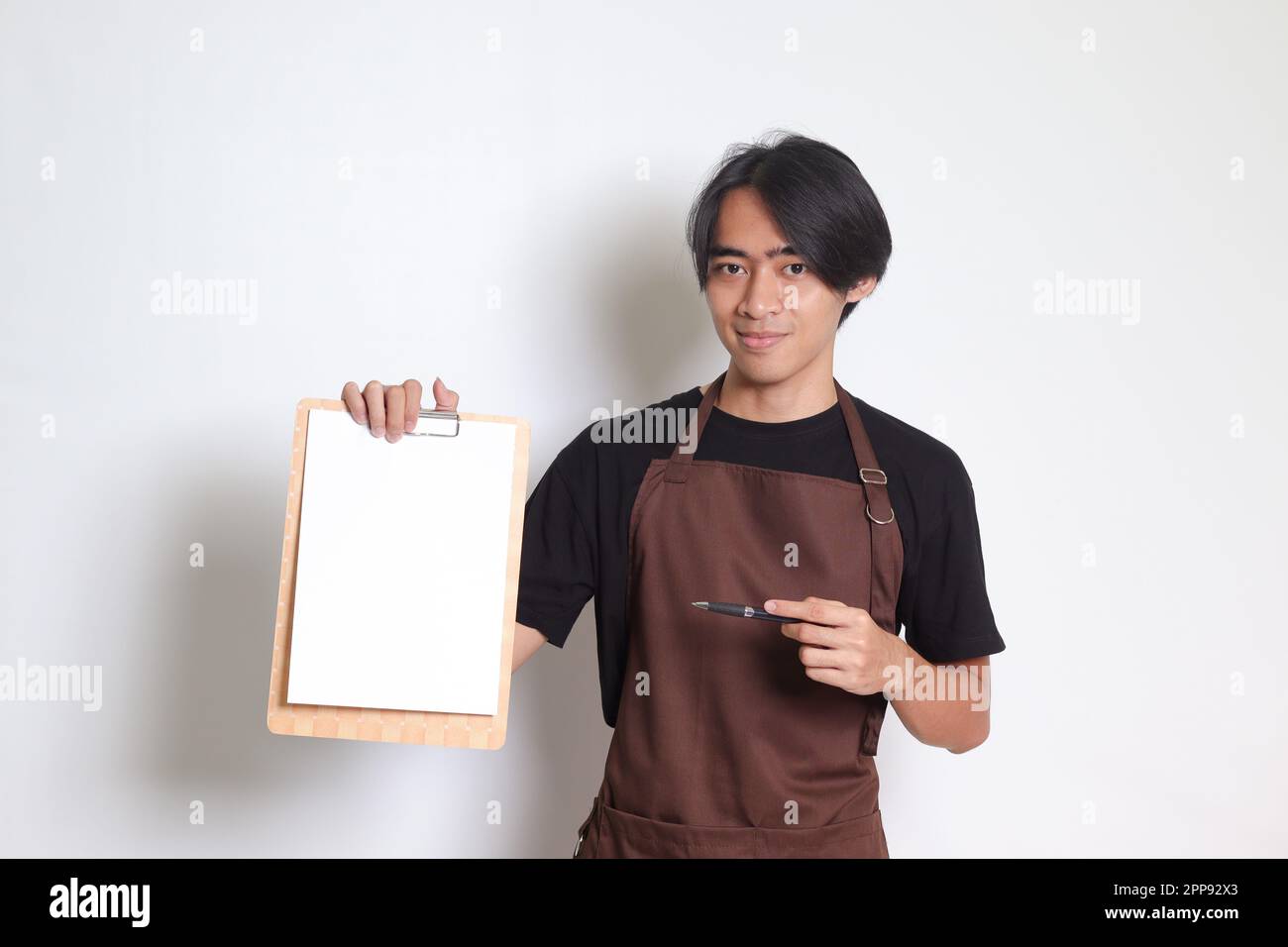 Ritratto dell'attraente barista asiatico in grembiule marrone con carta bianca del menu sulla clipboard. Immagine isolata su sfondo bianco Foto Stock