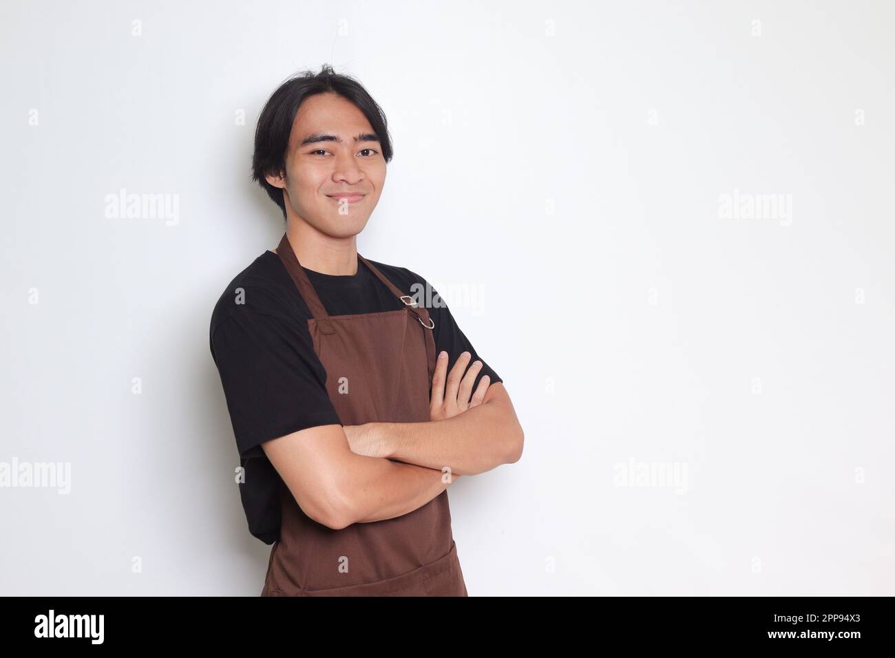 Ritratto di attraente barista asiatico uomo in grembiule marrone tenendo le braccia incrociate e guardando la fotocamera. Concetto pubblicitario. Immagine isolata su sfondo bianco Foto Stock