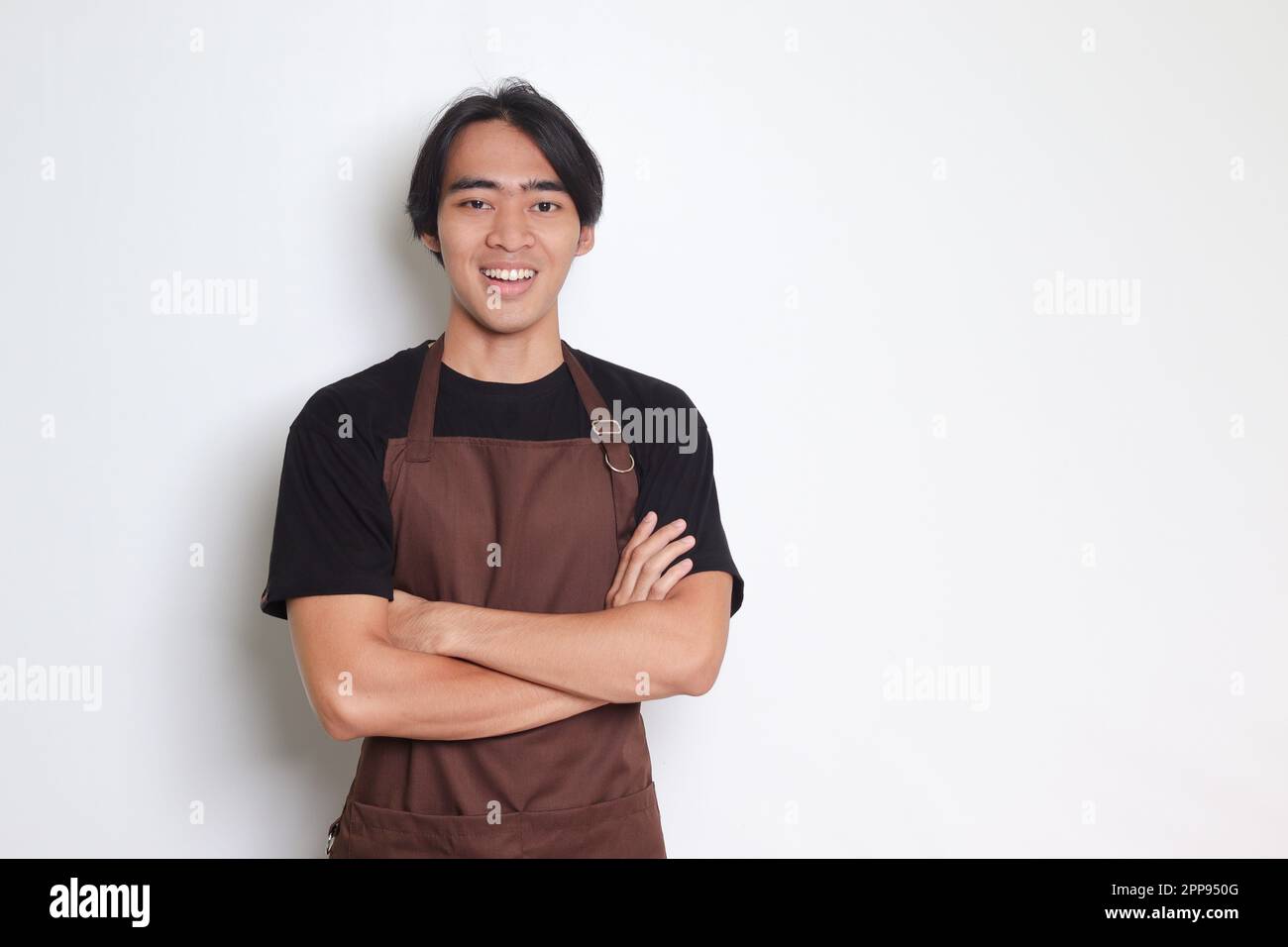 Ritratto di attraente barista asiatico uomo in grembiule marrone tenendo le braccia incrociate e guardando la fotocamera. Concetto pubblicitario. Immagine isolata su sfondo bianco Foto Stock