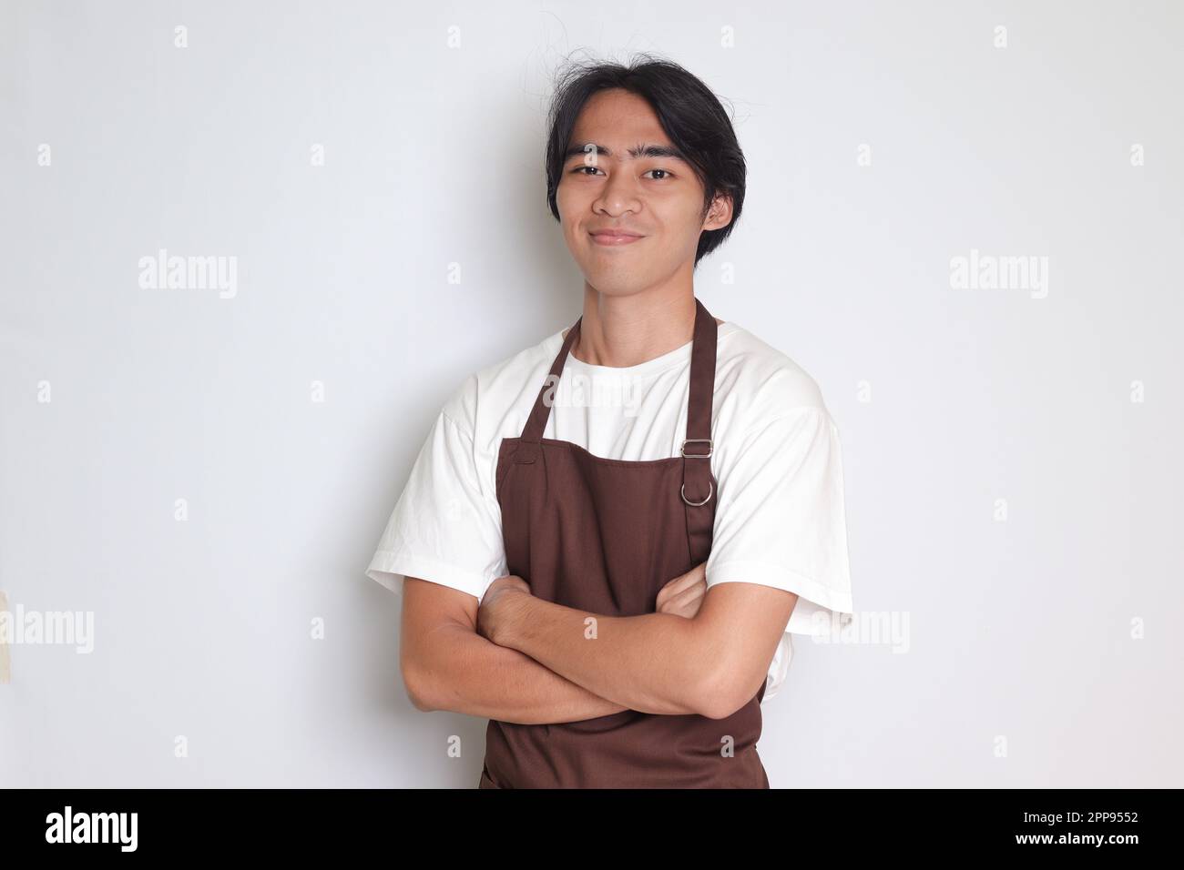 Ritratto di attraente barista asiatico uomo in grembiule marrone tenendo le braccia incrociate e guardando la fotocamera. Concetto pubblicitario. Immagine isolata su sfondo bianco Foto Stock