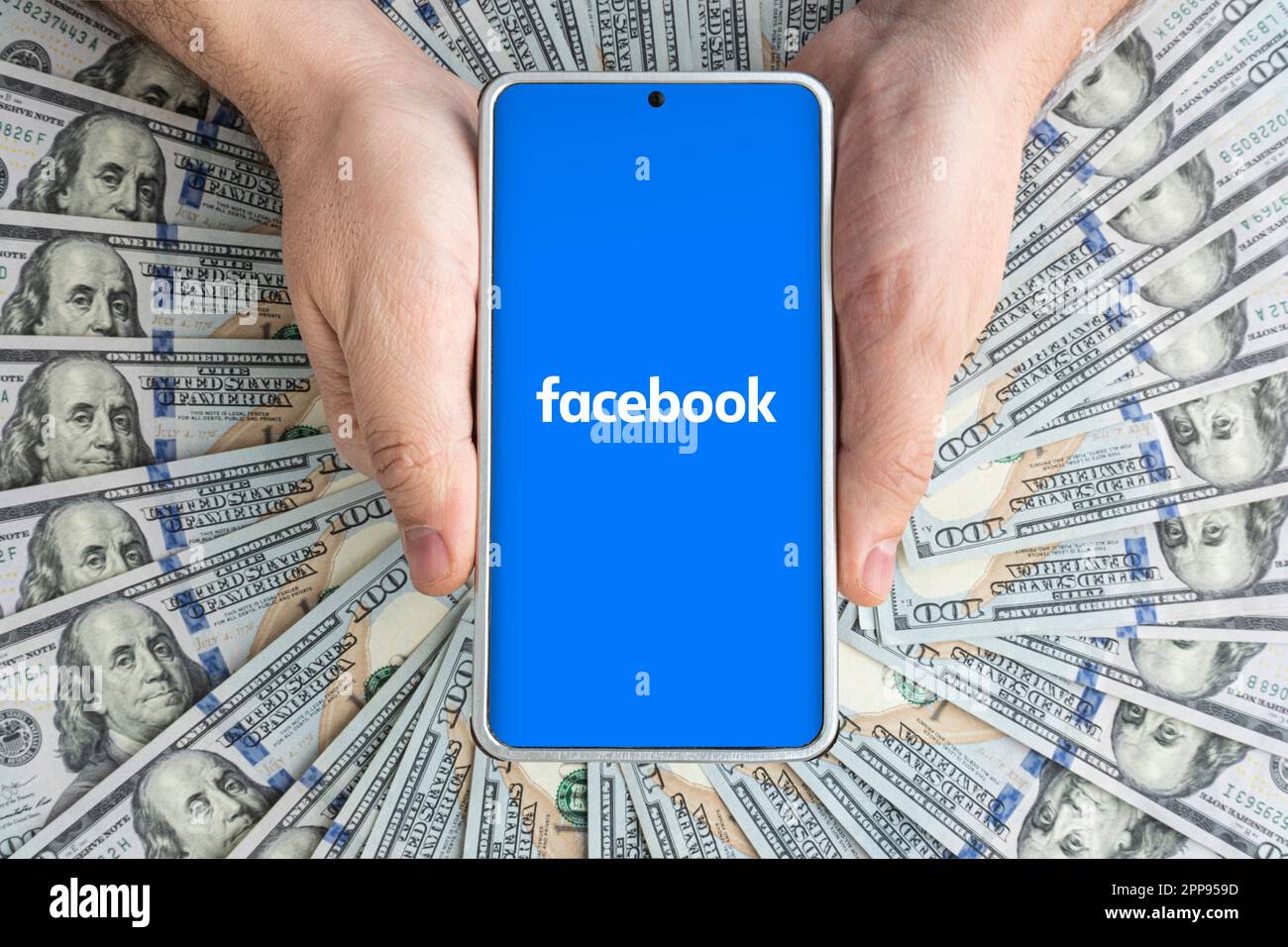 Schermo dello smartphone con l'app Facebook e molte banconote da cento dollari. Concetto di business e social networking. Marzo 30, 2023. Barnaul. Russia. Foto Stock