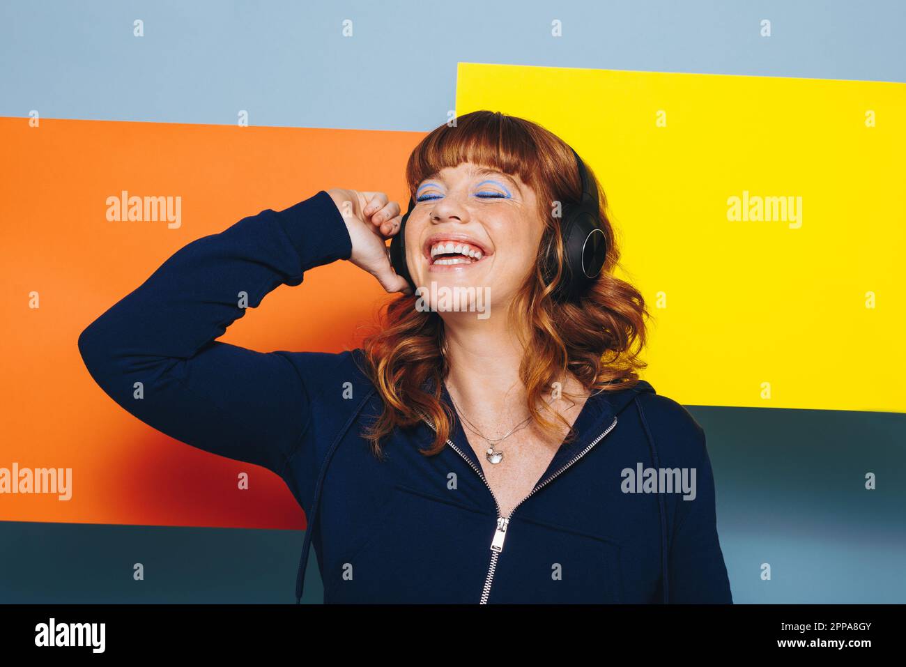 Donna che ascolta musica con cuffie wireless in uno studio di design. Felice giovane donna in piedi con forme geometriche colorate. Foto Stock