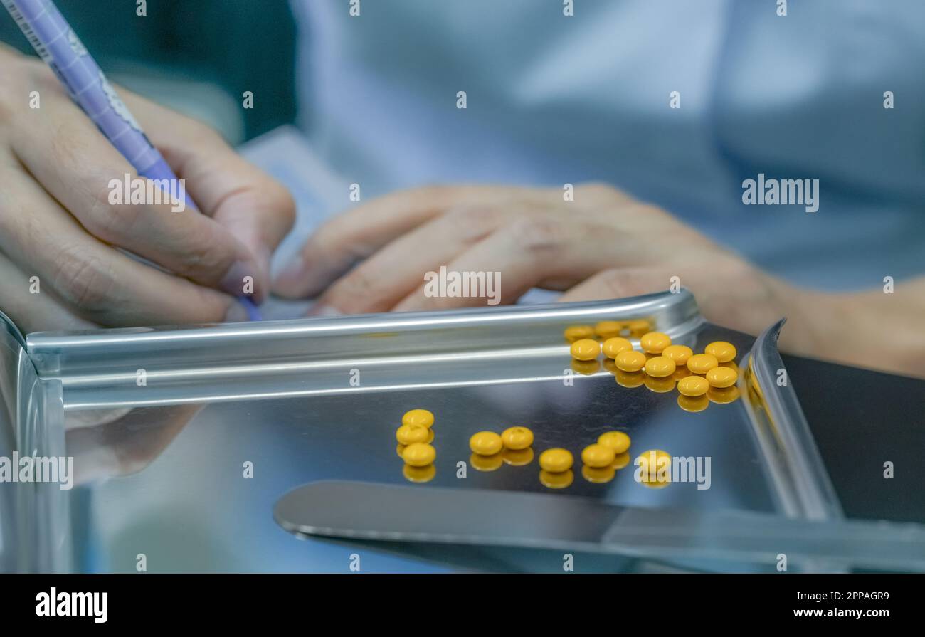 Compressa rivestita di zucchero giallo su vassoio di acciaio inossidabile Farmacista mano che tiene la penna e le istruzioni per la scrittura del farmaco sulla confezione di plastica della pillola. Pressione Foto Stock