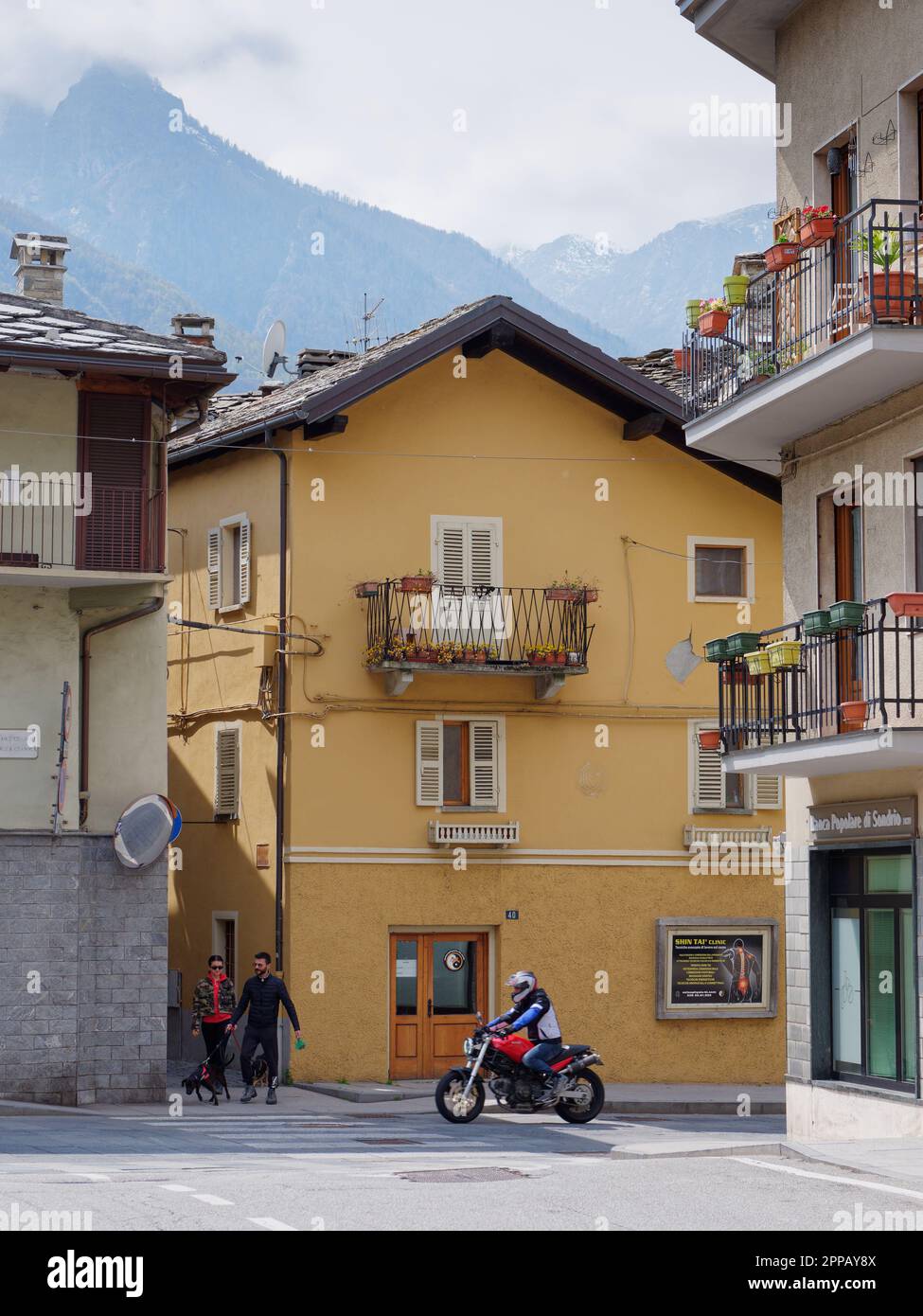 Pedoni e moto a Pont-Saint-Martin, Valle d'Aosta, NW Italia. Edificio con colorate fioriere su balcone e montagne sullo sfondo Foto Stock